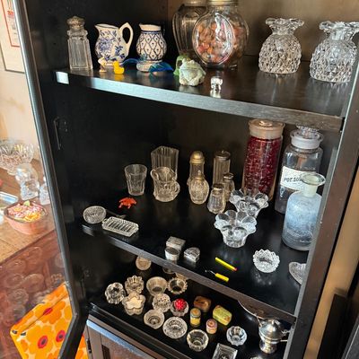 Sale Photo Thumbnail #180: Collection of vintage glassware, salt and pepper shakers, and small decorative trinkets displayed on the shelves of a dark wood cabinet.