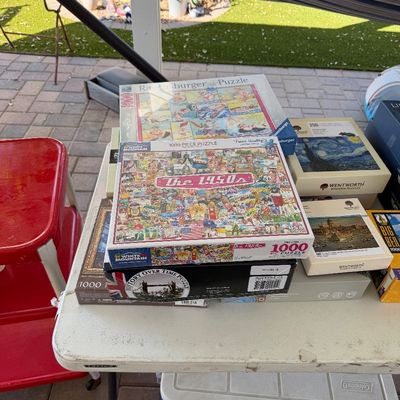 Sale Photo Thumbnail #158: Stack of assorted jigsaw puzzles, including a 1000-piece White Mountain "The 1950s" puzzle and a Ravensburger puzzle, displayed on an outdoor folding table.