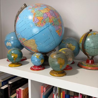 Sale Photo Thumbnail #35: Collection of vintage globes, including a large desk globe and several small tin lithograph globe banks, displayed on a white bookshelf.