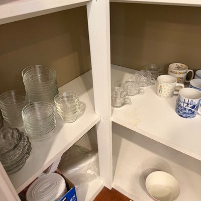 Sale Photo Thumbnail #150: Assorted glassware and ceramic mugs, including clear glass custard cups, patterned teacups, and a Beatrix Potter Peter Rabbit mug, displayed on white pantry shelves.