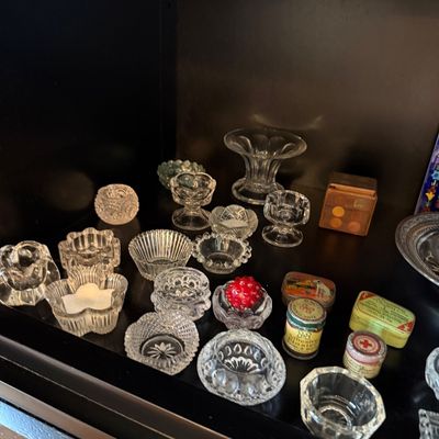Sale Photo Thumbnail #184: Collection of vintage pressed glass salt cellars in various patterns, displayed alongside small antique tins and a wooden box on a dark shelf.