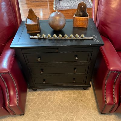 Sale Photo Thumbnail #214: Black three-drawer wooden end table flanked by red leather armchairs, displaying rustic decor including wooden wall pockets, a metal sphere, and a strap of brass sleigh bells.