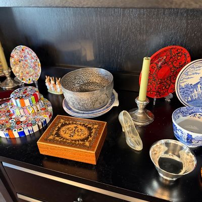 Sale Photo Thumbnail #192: Assorted vintage collectibles including a wooden inlaid music box, millefiori glass plates, an ornate silver-tone bowl, and a glass slipper figurine displayed on a dark wood cabinet.