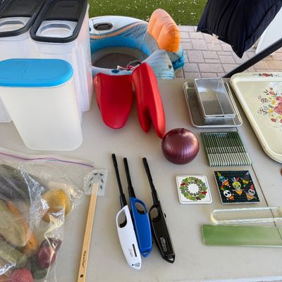 Sale Photo Thumbnail #153: Assorted household and kitchen items, including plastic storage containers, silicone oven mitts, utility lighters, coasters, and baking pans, displayed on a folding table.