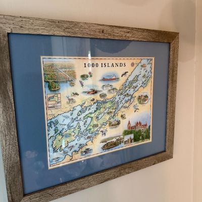 Sale Photo Thumbnail #174: Framed illustrated map of the 1000 Islands region, featuring local landmarks and wildlife, set in a blue mat and a rustic weathered wood frame.