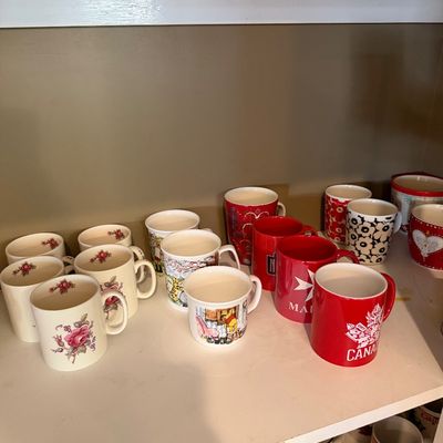 Sale Photo Thumbnail #151: Assortment of vintage and collectible ceramic coffee mugs, featuring floral patterns, souvenir designs, and novelty graphics, displayed on a white shelf.