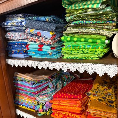 Sale Photo Thumbnail #66: Assorted stacks of colorful quilting and sewing fabric fat quarters, organized by color on wooden shelves with lace trim.