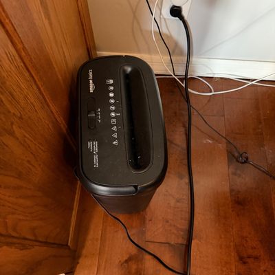 Sale Photo Thumbnail #204: Amazon Basics black paper shredder, showing light wear from use, plugged into a wall outlet on a hardwood floor.