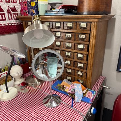 Sale Photo Thumbnail #53: Vintage oak multi-drawer library card catalog cabinet repurposed for craft storage, shown alongside a lighted makeup mirror and desk lamps on a red-and-white checkered tabletop.