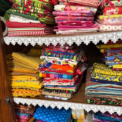 Sale Photo Thumbnail #67: Large collection of colorful quilting fabric fat quarters and remnants, neatly stacked by color and pattern on lace-trimmed wooden shelves.