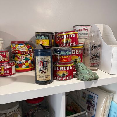 Sale Photo Thumbnail #39: Collection of vintage advertising tins, including Folger's Coffee, Chock Full o' Nuts, and Jaw Teasers, displayed alongside a Maytag oil can and a cast iron frog on a white shelf.