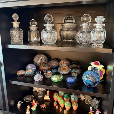 Sale Photo Thumbnail #190: Collection of vintage glass perfume bottles, millefiori paperweights, and miniature figurines displayed on black wooden shelves.