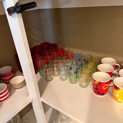 Sale Photo Thumbnail #149: Collection of vintage glassware and mugs, including floral-patterned tumblers in various colors and Marimekko-style bold floral mugs, displayed on white shelving.