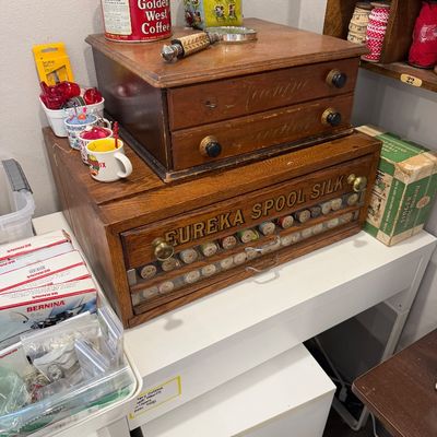 Sale Photo Thumbnail #51: Antique Eureka Spool Silk wooden cabinet with a glass-front drawer displaying vintage thread spools, topped with a smaller two-drawer wooden sewing chest.