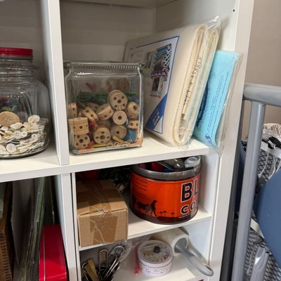 Sale Photo Thumbnail #42: Assorted crafting and sewing supplies, including a glass jar of wooden Tinker Toy pieces, a jar of vintage buttons, packaged batting, and a vintage Hills Bros. Coffee tin, organized on a white shelving unit.