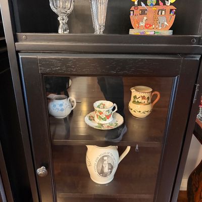 Sale Photo Thumbnail #196: Black wooden display cabinet with a glass door, showcasing a collection of vintage glassware, a Noah's Ark decorative piece, and various ceramic pitchers and teacups.