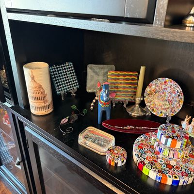 Sale Photo Thumbnail #194: Assorted decorative collectibles displayed on a black shelf, featuring colorful millefiori glass plates and stands, a blue Swedish Dala horse, and a cylindrical lamp depicting the US Capitol building.