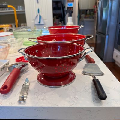 Sale Photo Thumbnail #133: Set of three red enamel colanders with silver-toned handles and rims, displayed alongside various kitchen utensils on a white countertop.
