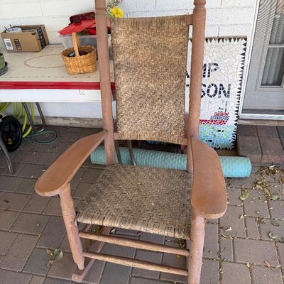 Sale Photo Thumbnail #164: Vintage wooden rocking chair with a woven splint seat and backrest, showing a weathered pinkish-brown painted finish.