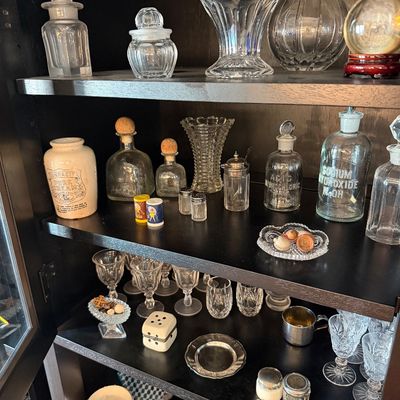 Sale Photo Thumbnail #195: Collection of vintage glassware, apothecary bottles, and small collectibles displayed on dark wooden shelves, including a Dunragit Pure Cream crock and labeled chemical bottles.