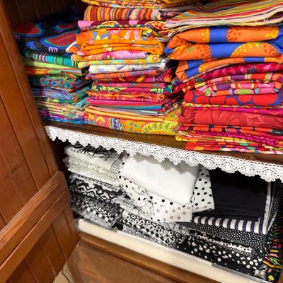 Sale Photo Thumbnail #65: Assorted stacks of sewing fabric, featuring vibrant multi-colored patterns on the top shelf and black-and-white prints on the bottom shelf, stored inside a wooden cabinet.