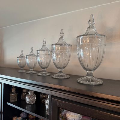 Sale Photo Thumbnail #201: Graduated set of five vintage clear glass apothecary jars with ribbed details and pointed finial lids, displayed atop a dark wood cabinet.