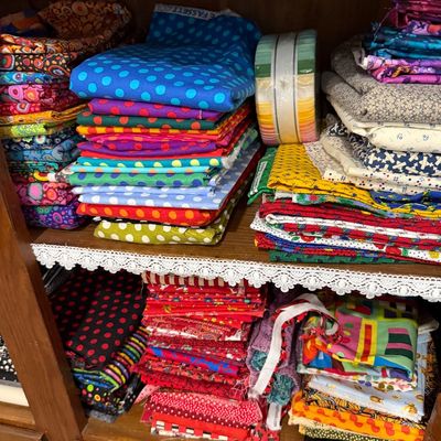 Sale Photo Thumbnail #62: Assorted quilting fabric quarters and fabric rolls, including Kaffe Fassett designs, organized on wooden shelves with lace trim.