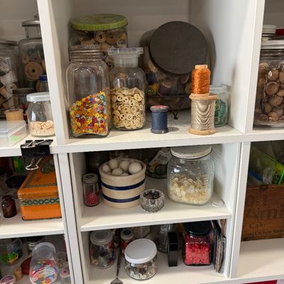 Sale Photo Thumbnail #38: Assorted vintage sewing notions, including buttons, thread spools, and beads, organized in glass jars on a white shelving unit.