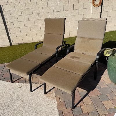 Sale Photo Thumbnail #159: Pair of outdoor chaise lounge chairs with black metal frames and tan cushions, showing some wear and weathering.