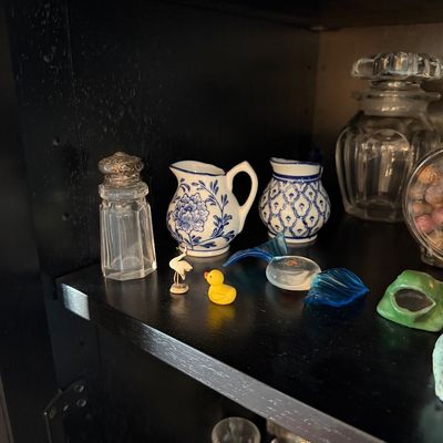 Sale Photo Thumbnail #181: Collection of miniature collectibles, including blue and white ceramic pitchers, a glass salt shaker with a metal lid, and small glass and plastic figurines displayed on a dark shelf.