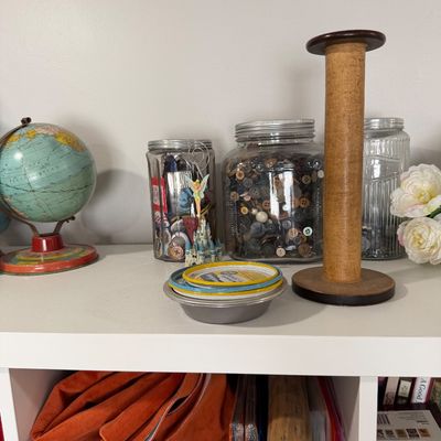 Sale Photo Thumbnail #32: Assorted vintage collectibles including a small tin globe, glass jars filled with buttons, and a tall wooden spool on a white shelf.