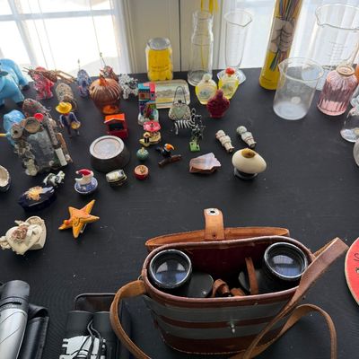 Sale Photo Thumbnail #88: Assortment of vintage collectibles, including binoculars in a leather case, small figurines, glassware, and decorative trinkets displayed on a black tabletop.
