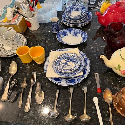 Sale Photo Thumbnail #143: Assorted vintage tableware collection, featuring blue and white transferware plates, yellow Fiestaware mugs, a red ceramic teapot, and various silver-plated serving spoons on a dark granite countertop.