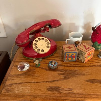 Sale Photo Thumbnail #3: Vintage red tin toy rotary telephone with paint wear, displayed alongside wooden alphabet blocks, a miniature rooster figurine, and other small collectibles on a wooden surface.