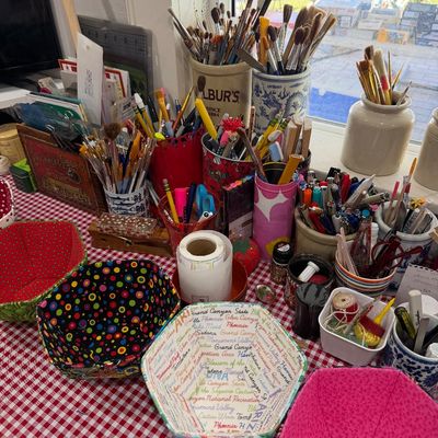 Sale Photo Thumbnail #57: Assorted craft and art supplies, including paintbrushes, pens, and pencils in various containers, alongside handmade fabric bowls on a red-and-white checkered tablecloth.
