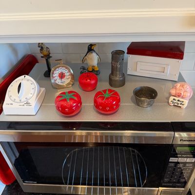 Sale Photo Thumbnail #113: Collection of vintage kitchen accessories and novelties, including Lux minute timers, tomato-shaped timers, a small scale, and decorative figurines, displayed atop a stainless steel Panasonic microwave.