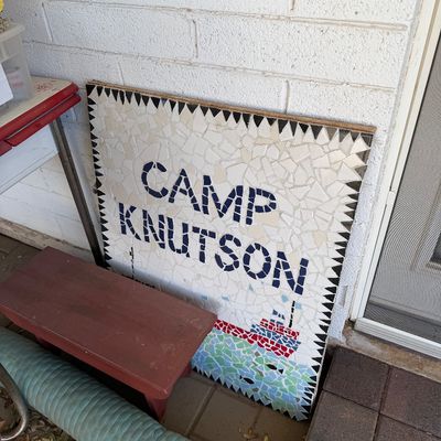 Sale Photo Thumbnail #169: Handmade mosaic sign reading "CAMP KNUTSON" with a sailboat design, featuring white, blue, red, and green tiles on a wooden backing.