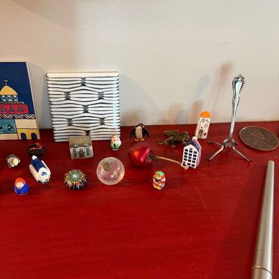 Sale Photo Thumbnail #79: Assortment of miniature collectibles, including small ceramic houses, a tiny penguin figurine, a painted rock, and a miniature fork stand, displayed on a red surface.