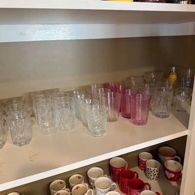 Sale Photo Thumbnail #148: Assorted glassware and ceramic mugs displayed on white shelves, including textured clear and pink tumblers and various patterned coffee cups.