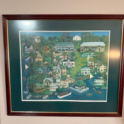 Sale Photo Thumbnail #2: Framed folk art style print depicting a lakeside community with Victorian cottages, boats, and figures, presented in a dark green mat and a reddish-brown wooden frame with a gold inner border.