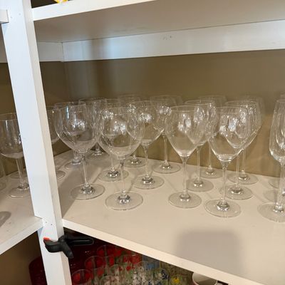 Sale Photo Thumbnail #147: Collection of clear glass wine goblets arranged on a white shelf, with various colorful drinking glasses and mugs visible on the shelf below.