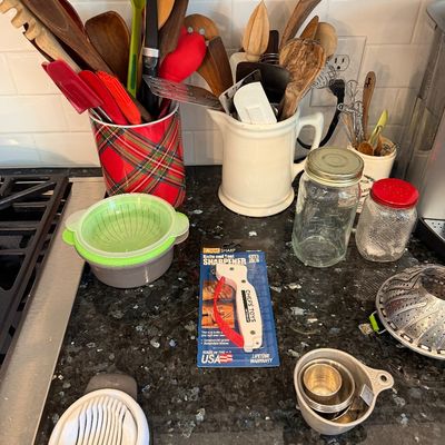 Sale Photo Thumbnail #121: Assorted kitchen utensils and gadgets, including wooden spoons in ceramic crocks, an egg slicer, a packaged knife sharpener, and glass jars on a dark granite countertop.