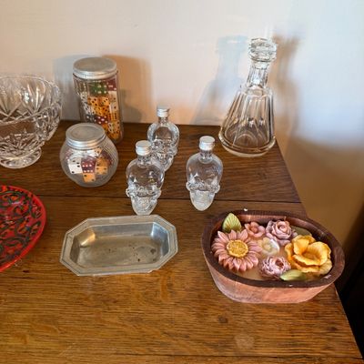 Sale Photo Thumbnail #173: Assorted tabletop decor including three glass skull bottles, jars filled with colorful dice, a crystal decanter, a pewter dish, and a wooden bowl with decorative wax flowers.
