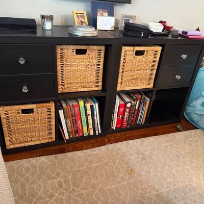 Sale Photo Thumbnail #83: Two dark brown IKEA Kallax shelving units arranged side-by-side, featuring drawer inserts with crystal-style knobs, woven wicker storage baskets, and books.