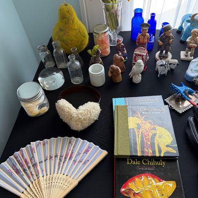 Sale Photo Thumbnail #96: Assortment of decorative items including Dale Chihuly art books, a cherry blossom folding fan, small glass bottles, and various figurines on a black tabletop.