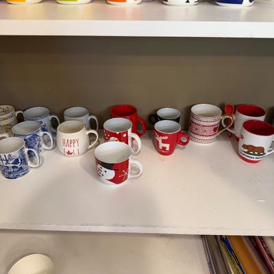 Sale Photo Thumbnail #141: Collection of assorted ceramic mugs on white shelves, featuring Marimekko Unikko floral mugs on the top shelf and various seasonal and novelty mugs below.