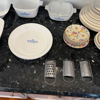 Sale Photo Thumbnail #139: Vintage Corning Ware Blue Cornflower casserole dishes and pie plates, shown alongside floral patterned plates and three metal hand graters on a dark countertop.