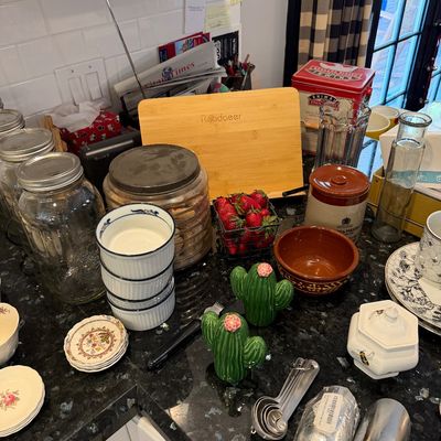 Sale Photo Thumbnail #144: Assorted kitchenware including a bamboo cutting board, glass storage jars, ceramic bowls, cactus-shaped salt and pepper shakers, and metal measuring spoons on a dark granite countertop.