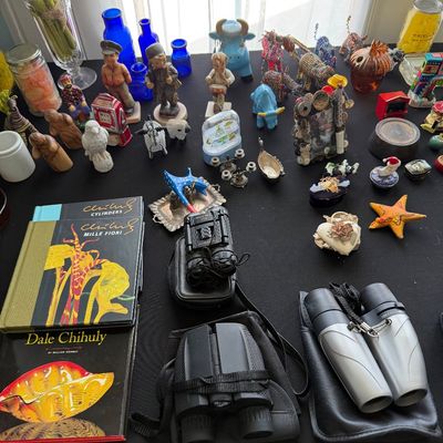 Sale Photo Thumbnail #87: Assortment of collectible figurines, blue glassware, Dale Chihuly art books, and multiple pairs of binoculars displayed on a black tabletop.