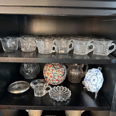 Sale Photo Thumbnail #198: Collection of vintage glassware and ceramics, including clear pressed glass punch cups, a colorful floral ceramic sphere, a small blue and white porcelain plate, and a floral teapot, displayed on black shelves.